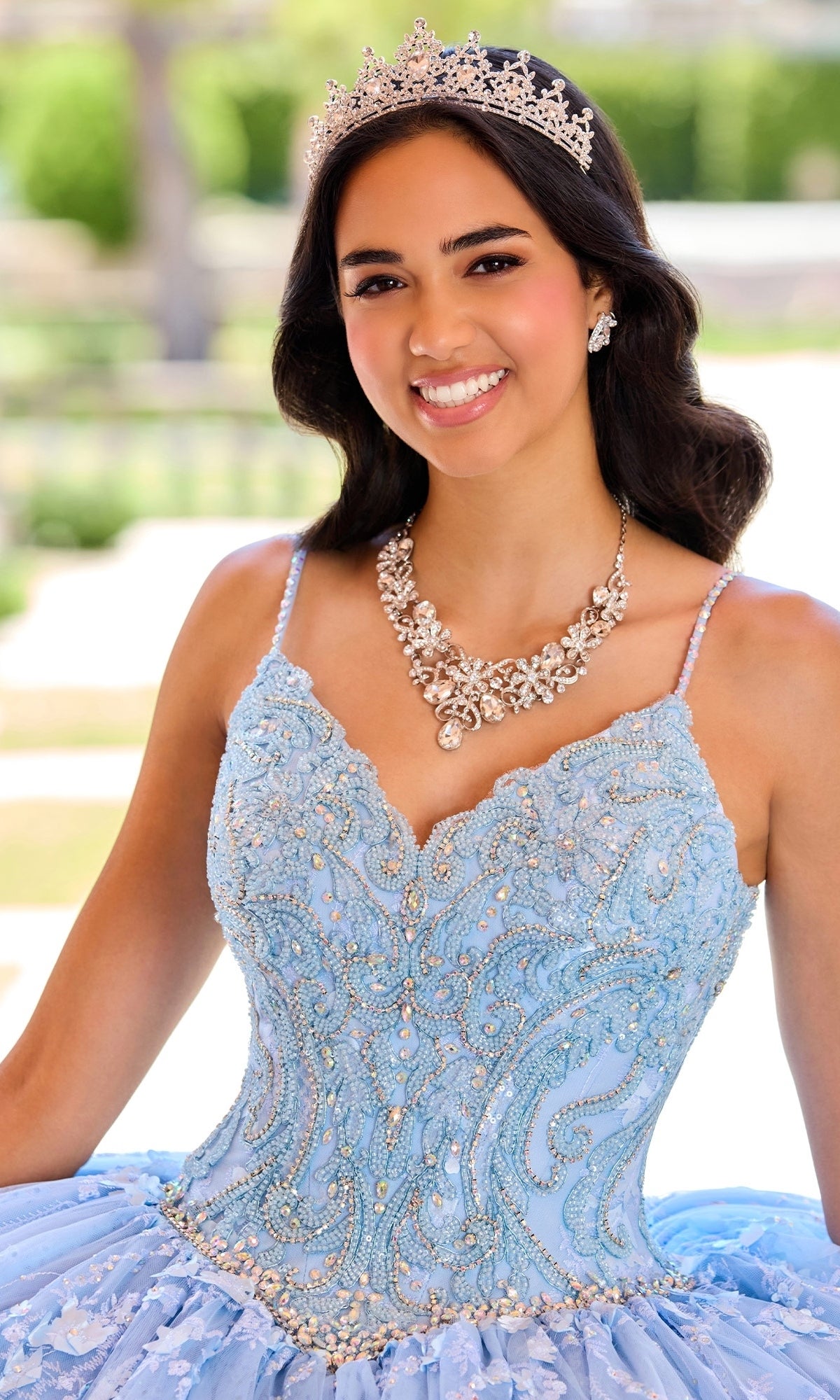 A young woman smiles outdoors wearing a jeweled tiara, ornate necklace, and the Princesa by Ariana Vara Lace Quince Dress PR12268 with delicate beading.